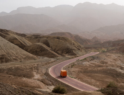 View of a vibrant orange vehicle winds its way through the undulating ochre and tan terrain under a hazy sky, Zhangye, Gansu, China.
