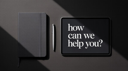 Top view black office desk with notebook pen and tablet displaying "how can we help you?" text