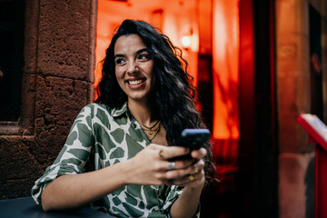Obraz premium Smiling young woman holds smartphone while leaning on table outside café, radiating positive energy, comfort and confidence in a relaxed moment of digital connectivity.