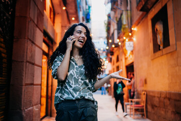 Obraz premium Excited woman gestures mid-call with expressive face, talking via smartphone in city alley, conveying engagement, connection and digital immersion in modern lifestyle.