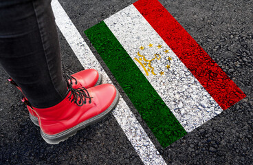 a woman with a boots standing on asphalt next to flag of Tajikistan and border

