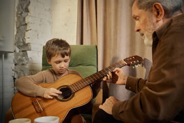 Senior mentor showing guitar chords to young boy at home. Concept of step by step learning, music instruction, family mentoring, creative education, skill transfer.
