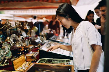 Asian woman joyfully explores vintage jewelry at flea market, smiling with delight, expressing fascination, curiosity and appreciation for cultural objects and timeless aesthetics.