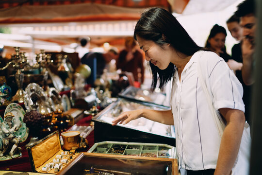 Asian woman joyfully explores vintage jewelry at flea market, smiling with delight, expressing fascination, curiosity and appreciation for cultural objects and timeless aesthetics.