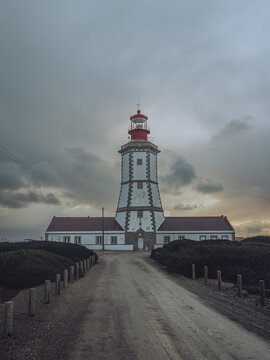View of a towering lighthouse with a red cap piercing the stormy sky, standing sentinel on the rugged coastline, Farol do Cabo Espichel, Setubal, Portugal.