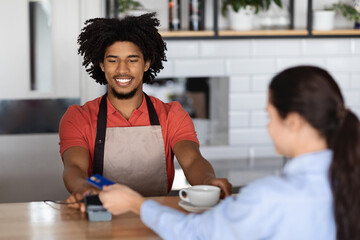 Smiling curly attractive young black man bartender in apron at bar counter gives terminal to lady...