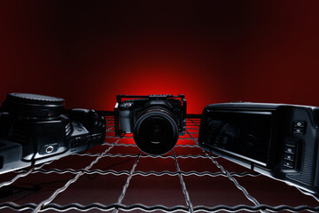 A camera and two other gadgets rest on a grid pattern with a dark red background. The arrangement highlights tools often used in photography and videography settings.