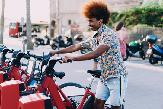 Smiling young man with curly afro unlocking rental bike at city dock. Conceptual lifestyle photo representing app-based mobility, urban technology, and eco-friendly commuting. - Powered by Adobe