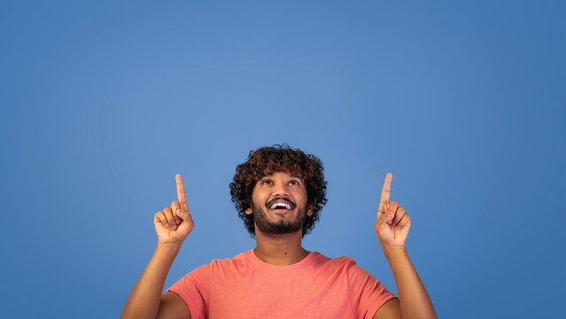 A young man with curly hair smiles widely while pointing both of his index fingers up. He wears a light orange shirt. The background is a solid blue color.