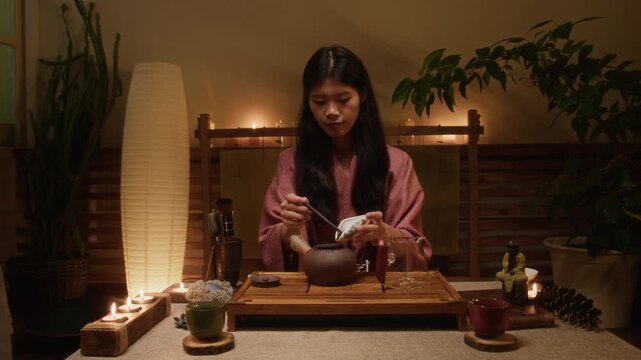 Medium shot of calm, relaxed young Chinese woman, dressed in pink hanfu kimono, sitting on floor at table at traditional home, smelling dry leaves then placing in clay teapot to brew tea gongfu style