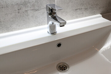Fototapeta premium sleek chrome single-handle faucet mounted on a white ceramic sink. Water flows smoothly from the spout; the sink features a central drain hole. Set against a textured gray wall