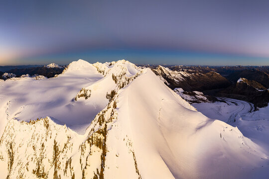 Aerial view of snow-capped peaks pierce the sky as dawn paints the horizon with hues of rose and gold, casting long shadows across the pristine landscape, Piz Palu, Pontresina, Grisons, Switzerland.