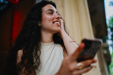 Smiling woman with curly hair holding smartphone, embodying joy and connection in a mobile-first...