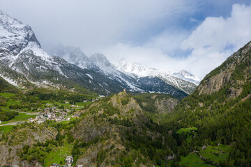 Fototapeta premium A traditional alpine village and a historic stone tower perch on green hillsides beneath snow-capped peaks in Val d'Aoste, Italy. The landscape features dramatic mountain slopes, dense forests, and