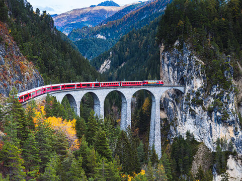 Aerial view of a vibrant red train gracefully traverses the Landwasser Viaduct amidst the autumn foliage, Bergun Filisur, Grisons, Switzerland.