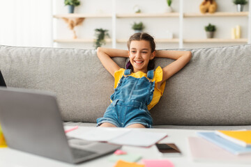 Rest Concept. Smiling female teenager sitting on comfortable couch, leaning back on sofa and looking at laptop screen, watching movie or video, taking break from studying online at home
