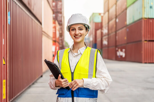 Smiling female logistics manager in hardhat and safety vest holding tablet, standing at shipping container terminal port. Professional warehouse inspector overseeing global export import industry.