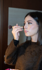 Young woman in sweater applies natural makeup with a brush, profile close-up capturing skincare and grooming routine for a fresh, confident morning look in soft indoor light