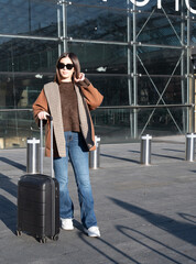 Young woman standing outdoors in front of a contemporary glass building, holding a black rolling suitcase, ready for a journey or commute in an urban setting