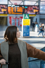 Young woman standing in bright airport terminal with luggage, looking toward departures board through large windows, poised and ready for an international journey or business trip