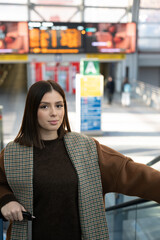 Young woman standing on an escalator holding a luggage handle, preparing for a professional or personal journey inside a modern transportation hub with a departure board in the background