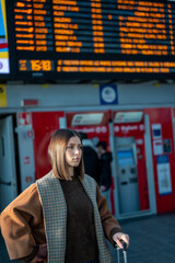 Woman standing in a train station, pulling a suitcase while checking the departures board, representing themes of travel, commuting, and urban transportation awaiting a journey