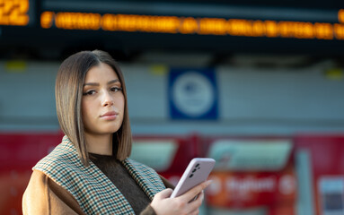 Young woman standing on a train station platform, holding her mobile phone and waiting for public transport, with a blurred information board in the background