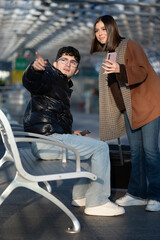 Young man sitting on a bench pointing finger while a woman stands next to him holding a smartphone, both appearing to navigate or seek information at a transit station