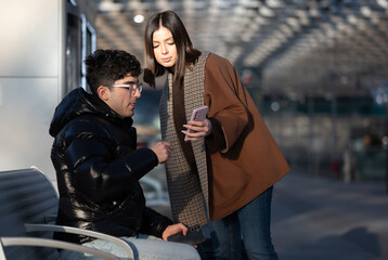Young couple comparing information on their smartphones, sharing connection and digital media while waiting in a modern public transport station, representing modern travel and communication