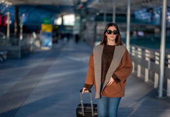 Young woman wearing a brown coat and sunglasses walking confidently with her suitcase through a modern airport terminal, ready for travel and a journey vacation