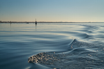 Waves ripple across sunlit water, created by a passing vessel, near buoys, medium shot