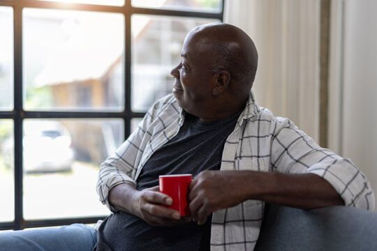 Senior man sits comfortably at home, holding a red mug and gazing thoughtfully out the window, enjoying a quiet moment of reflection.