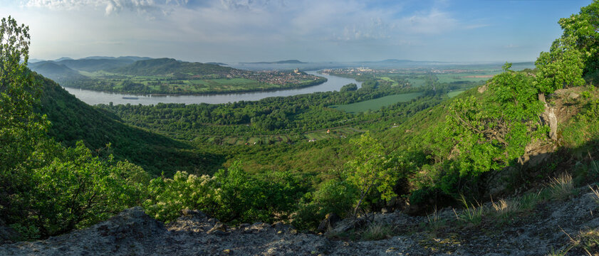 View of a wide river snaking through lush green valleys and hills under a soft sky, cutting through a distant town, Sturovo, Nitra Region, Slovakia.