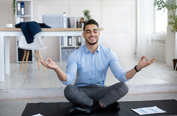 Workplace stress management concept. Calm Arab man meditating with closed eyes at modern office. Young businessman feeling peaceful and balanced, doing yoga on mat