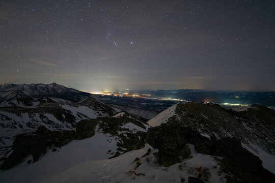 View of snow-dusted peaks under a starlit sky, with a distant city's lights shimmering like jewels on the horizon, Nizna Bystra, Zilina Region, Slovakia.