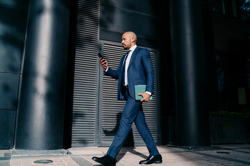 Focused businessman in tailored suit walking confidently in urban business district, multitasking with smartphone and digital tablet, symbolizing mobile decision-making and digital leadership.