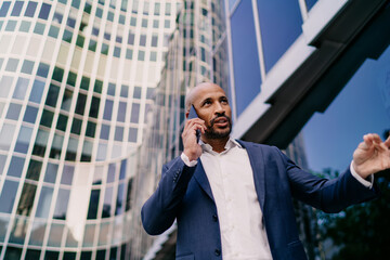 Focused businessman gesturing while on phone under mirrored skyscraper, illustrating negotiation, leadership and digital decision-making in high-pressure business setting.