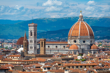 Panoramic view of Florence (Firenze), Italy, showing rooftops, landmarks like Palazzo Vecchio and Santa Maria del Fiore with its red dome, and hills with forests on a sunny summer day.