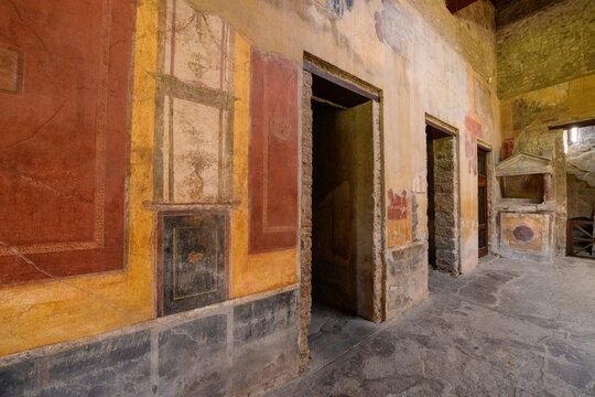 Ancient Roman hallway with vibrant wall paintings and textured stone floors in the House of Menander, Pompeii, Italy. Faded frescoes in ochre, red, and yellow tones frame doorways and highlight the