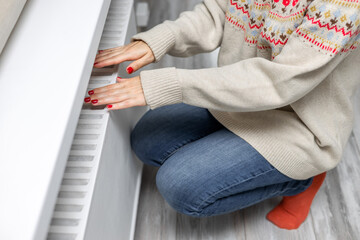 A girl in a knitted sweater warms her hands near a radiator. A girl warms herself near a heating radiator. heating radiator. heating season. 