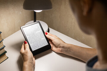 A woman reads an e-reader by the light of a table lamp. A woman reading an e-book at home. reading a book at the table. 