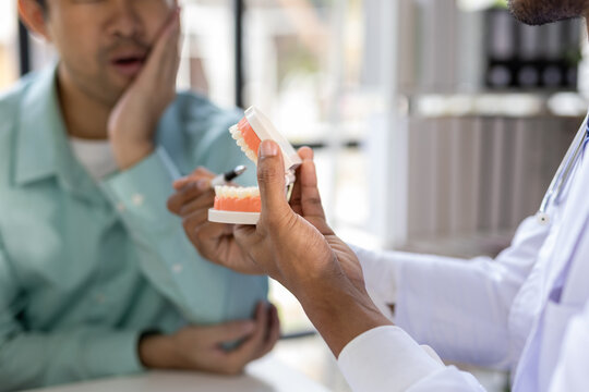 Man with toothache consults with doctor in examination room. Dentist demonstrating an anatomical model of the human jaw while explaining treatment options to a patient in a dental clinic.