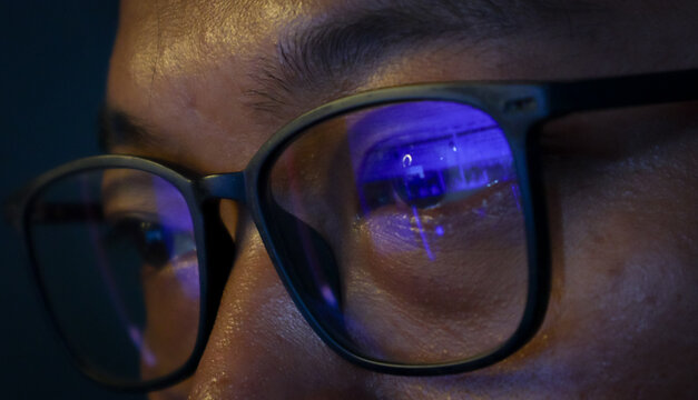 Close up view of focused businessman wears computer glasses for reducing eye strain blurred vision looking at pc screen with computer reflection using internet, reading, watching, working online late.