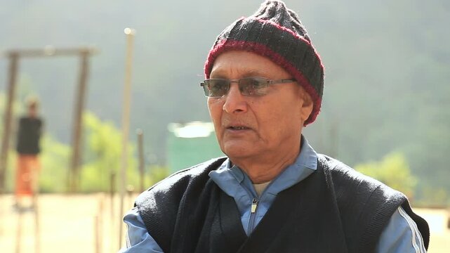 An elderly man around 70 years old sits outdoors talking to someone, reflecting rural life, experience, and peaceful daily moments in a mountain village of Uttarakhand, India.