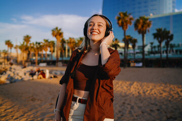 Fototapeta premium Young woman listening to music on beach with headphones, smiling at camera, representing connection between joy, tech and daily escape through immersive digital experiences.