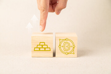 A hand points to wooden blocks featuring gold bars and Bitcoin icons with upward arrows, symbolizing financial growth, investment strategy, and choosing between digital assets and traditional gold.