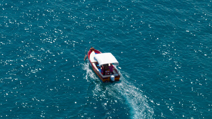 High-angle shot of a small wooden boat with a white canopy cruising through sparkling turquoise water, leaving a gentle white wake on the sun-dappled sea surface. Aerial view of a boat in the sea.