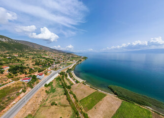High-angle aerial panorama of the Lake Ohrid coastline near Udenisht, Albania, featuring the turquoise lake waters, coastal road, rural farmlands, and rolling hills under a blue sky.