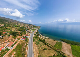 High-angle aerial panorama of the Lake Ohrid coastline near Udenisht, Albania, featuring the turquoise lake waters, coastal road, rural farmlands, and rolling hills under a blue sky.