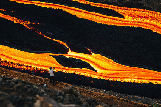 View of fiery rivers of molten lava snake through the dark, rugged landscape as a lone photographer captures the raw power, Fagradalsfjall, Reykjanes Peninsula, Iceland.